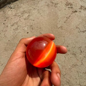 Puede incluir: Un objeto esférico liso de color rojo profundo translúcido, que presenta una banda naranja brillante distintiva que recorre diagonalmente su superficie, creando un contraste vibrante contra los tonos rojos más oscuros de la esfera pulida.