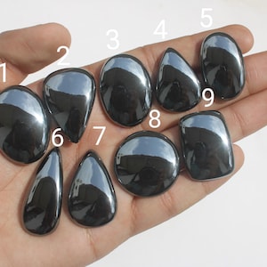 May include: Nine polished, dark gray, metallic hematite cabochons in various shapes including oval, teardrop, and round. Each stone is numbered from one to nine in white text, showcasing a smooth, reflective surface and a high-gloss finish.