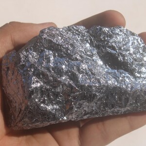 May include: A raw, jagged chunk of metallic silicon with a highly reflective, silver-grey surface. The mineral specimen features sharp, crystalline edges and a complex, multi-faceted texture that catches light across its irregular, brittle-looking form.
