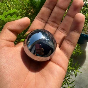 Puede incluir: Un objeto esférico liso y pulido con una superficie metálica oscura altamente reflectante. La esfera actúa como un espejo, mostrando el reflejo claro de una persona sosteniendo un teléfono inteligente mientras captura la imagen con luz natural.