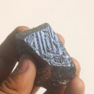 May include: A raw, dark grey silicon crystal specimen with a metallic lustre and a distinct, geometric, grid-like crystalline structure on its surface. The mineral fragment features sharp, angular edges and a reflective, multifaceted texture.