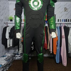 Puede incluir: Un disfraz de cuerpo completo de Green Lantern que presenta un mono negro con detalles verdes en hombros, antebrazos y rodillas. El pecho muestra un emblema circular verde y blanco. El traje incluye secciones acolchadas en el torso.