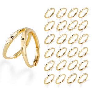 Op de afbeelding: Een collectie van goudkleurige metalen ringen, elk voorzien van een enkele gegraveerde letter van het alfabet. Twee ringen worden op de voorgrond getoond met de letters A en B, terwijl de overige ringen van C tot Z in een raster worden weergegeven.