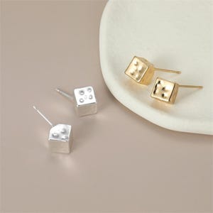 May include: A pair of gold-toned cube-shaped stud earrings and a pair of silver-toned cube-shaped stud earrings. Each earring features a smooth, metallic finish with small, indented circular markings on the top surface, resembling the face of a die.