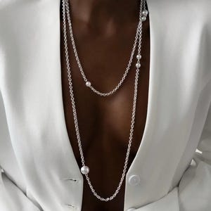 Può includere: Una lunga collana a catena color argento con diverse perle sferiche bianche di varie dimensioni distanziate lungo le maglie. La collana è indossata sopra un blazer bianco con un unico bottone visibile, creando un aspetto a strati.