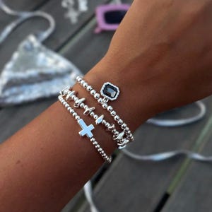 May include: A set of three silver-toned beaded bracelets. One bracelet features a small silver cross charm, another has a series of textured metallic accents, and the third includes a rectangular dark gray gemstone charm with a silver border.