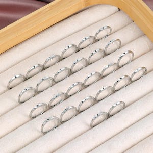 Peut inclure: Une collection de bagues fines en métal argenté, chacune gravée d'une lettre unique de l'alphabet de A à Z. Les bagues sont disposées en rangées sur un plateau de présentation rembourré de couleur claire avec un cadre en bois.