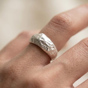 Op de afbeelding: Een zilveren ring met een dikke, afgeronde band met een sterk getextureerd, onregelmatig oppervlak dat het uiterlijk van ruw, gehamerd metaal nabootst. De afwerking is reflecterend en helder, met diepe inkepingen en ribbels over de buitenkant.
