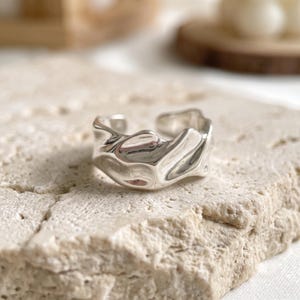 May include: A silver-toned ring with an irregular, molten-like surface texture. The band features a wide, open design with a polished, reflective finish that catches the light, creating deep shadows and bright highlights across its organic, wavy form.