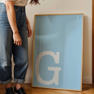 Op de afbeelding: Een grote rechthoekige wandposter met een lichtblauwe achtergrond en een opvallende, grote witte hoofdletter G in het midden. De poster is ingelijst in een dunne, lichtgekleurde houten lijst en staat rechtop op de vloer.