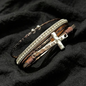 Op de afbeelding: Een armband met meerdere strengen, bestaande uit een dun bruin koord met twee zilverkleurige kralen, een band bezet met heldere strass-steentjes en een centraal zilverkleurig metalen kruis met de inscriptie BLESSED, vastgezet met bruine koordjes.