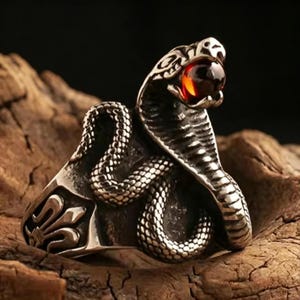 Puede incluir: Un anillo de metal plateado con forma de serpiente cobra con cuerpo enroscado y escamas texturizadas. La cabeza de la serpiente está levantada y sostiene una pequeña piedra preciosa redonda, translúcida y de color naranja oscuro.