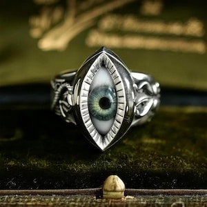 May include: A silver ring featuring a central marquise-shaped setting that contains a realistic, detailed human eye with a green iris and black pupil. The metal band is decorated with intricate, raised scrollwork patterns along the sides.