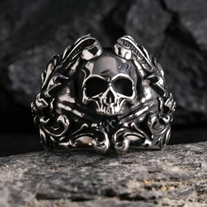Könnte beinhalten: Ein silberfarbener Metallring mit einem zentralen Totenkopfmotiv, das von kunstvollen, erhabenen Schnörkeln und blattartigen Mustern umgeben ist. Die Oberfläche des Rings ist poliert und weist dunkle Vertiefungen auf, die die Details betonen.