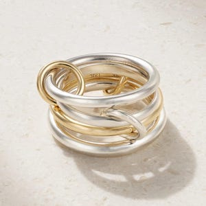 May include: A multi-band ring featuring interlocking silver and gold-toned circular bands. The interior of the gold band is clearly engraved with the text 18K, while the silver band features an engraved hallmark. The design consists of several smooth, rounded metal loops that overlap to create a layered, cohesive structure.