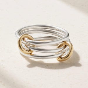 May include: A multi-band ring featuring three smooth silver-toned circular bands that are interconnected by two smaller, polished gold-toned oval links. The design creates a layered, continuous loop effect with a polished metallic finish throughout.