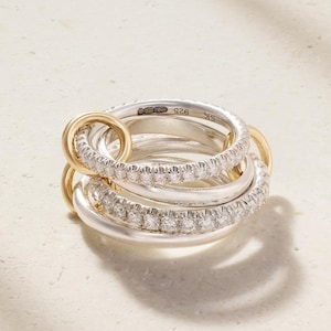 May include: A multi-band ring featuring interlocking silver and gold-toned circles. Two of the silver bands are pavé-set with small clear stones. The interior of one silver band is engraved with the text 925 and SK, indicating sterling silver.