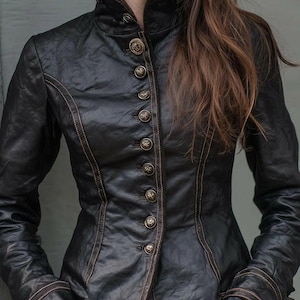 May include: A black leather jacket featuring a high collar, long sleeves, and a front button closure with ornate metallic buttons. The jacket is detailed with visible brown stitching along the seams and cuffs, creating a structured silhouette.