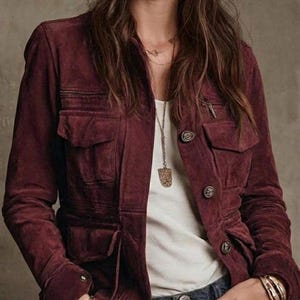 May include: A deep burgundy suede jacket featuring a collared neckline, long sleeves, and four front flap pockets. The jacket is styled with a white undershirt, a long pendant necklace, and blue denim jeans, showcasing a button-front closure.