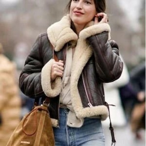 Könnte beinhalten: Eine dunkelbraune Lederjacke mit einem dicken, cremefarbenen Lammfellkragen und Ärmelbündchen, mit silbernem Reißverschluss, getragen über einem hellbeigen Oberteil und einer blauen Jeans, kombiniert mit einer beigen Wildleder-Beuteltasche.