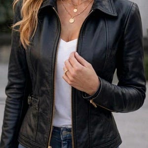 May include: A black leather jacket featuring a pointed collar, long sleeves, and a gold-toned front zip fastening. The jacket includes vertical seam detailing along the front panels and a small side buckle adjustment at the waistline.