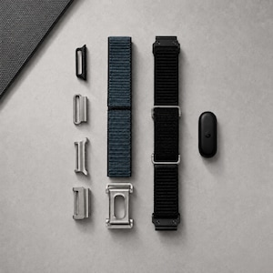 May include: A collection of watch accessories including a dark blue fabric strap, a black fabric strap with silver-toned metal hardware, four individual silver-toned metal connector pieces, and a small black oval-shaped electronic device component.