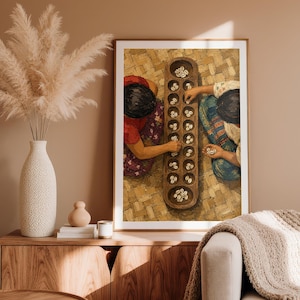 May include: A framed wall art print depicting two individuals playing a traditional board game with small white shells on a wooden board. The scene shows a top-down view of the players, their colorful clothing, and the textured woven mat surface.