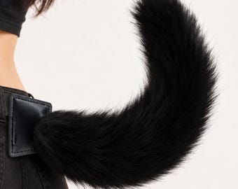 May include: A long, fluffy black faux fur tail attached to a black rectangular leather-like patch. The patch is secured to the waistband of black denim jeans. The tail curves upward in a sweeping motion, displaying dense, dark fibers.