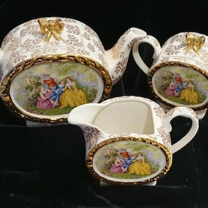 May include: A porcelain tea set comprising a teapot, a lidded sugar bowl, and a creamer. Each piece features a white base with gold leaf patterns, gold-trimmed oval medallions, and a central colourful scene of figures in a garden setting.