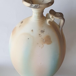 May include: A ceramic pitcher with a rounded body and a flared rim. The surface features a matte finish with blended gradients of cream, soft blue, and pink, accented by dark brown mottled spots and a single curved handle on the side.