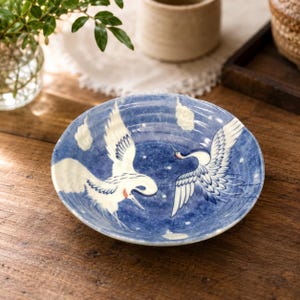 May include: A round ceramic bowl featuring a deep blue mottled background with two white cranes in flight. The design includes small white clouds and subtle white dots scattered across the surface, with the cranes displaying blue wing details.