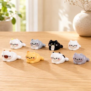 May include: A collection of eight small, smooth, ceramic cat figurines in various colors including white, gray, black, and yellow. Each cat is depicted in a resting, prone position with simple, stylized facial features, including black eyes and pink cheeks.
