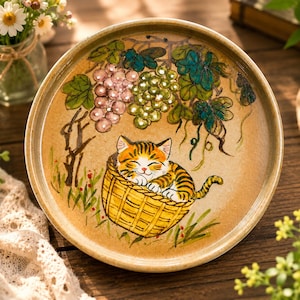 May include: A round ceramic plate with a tan speckled finish featuring a hand-painted illustration of a ginger tabby kitten sleeping inside a yellow woven basket, positioned beneath hanging clusters of purple and green grapes with dark green leaves.