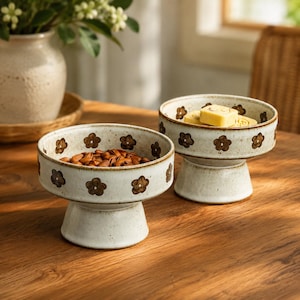 May include: Two off-white ceramic pedestal bowls with a speckled finish and a repeating pattern of small, dark brown hand-painted flowers. One bowl contains a pile of brown almonds, while the other holds two square, light yellow food items.