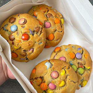 May include: Three round, golden-brown baked biscuits nestled inside a white cardboard box lined with white parchment paper. Each biscuit is studded with numerous multi-coloured candy-coated chocolate pieces in shades of red, blue, orange, yellow, pink, and purple.