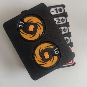 May include: A black plastic game counter featuring two circular orange dials with the numbers 11 and 10 printed on them. Beneath the main housing, several white and red rectangular tabs are visible, each marked with numbers and a small leaf icon.