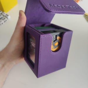 May include: A purple deck box with a textured exterior and a hinged lid featuring the embossed text Mage Tech. The box has a front cutout revealing a black and orange circular emblem on a card inside, with a clear window on the side panel.