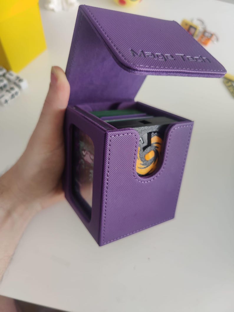May include: A purple deck box with a textured exterior and a hinged lid featuring the embossed text Mage Tech. The box has a front cutout revealing a black and orange circular emblem on a card inside, with a clear window on the side panel.