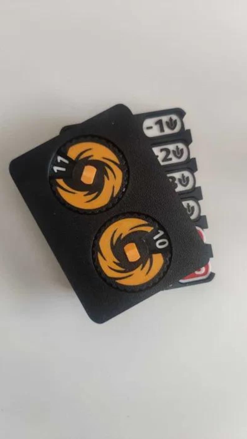 May include: A black plastic game counter featuring two circular orange dials with the numbers 11 and 10 printed on them. Beneath the main housing, several white and red rectangular tabs are visible, each marked with numbers and a small leaf icon.