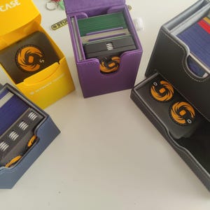 May include: Four card deck cases in yellow, purple, blue, and gray. The yellow case is open, revealing a black box with an orange circular logo. The purple and blue cases hold card stacks and tokens. The gray case features a drawer with two orange tokens.