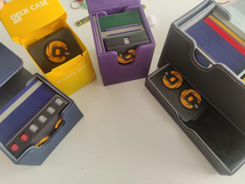 May include: Four card deck cases in yellow, purple, blue, and gray. The yellow case is open, revealing a black box with an orange circular logo. The purple and blue cases hold card stacks and tokens. The gray case features a drawer with two orange tokens.