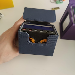 May include: A navy blue textured deck box with an open lid, revealing a stack of playing cards inside. The cards feature a black divider with a graphic of two orange eyes. The box has a rectangular cutout on the front and stitched edges.