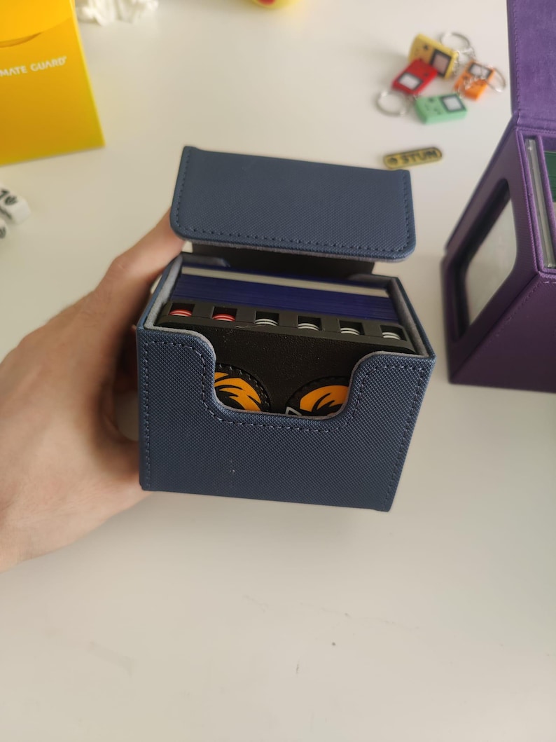 May include: A navy blue textured deck box with an open lid, revealing a stack of playing cards inside. The cards feature a black divider with a graphic of two orange eyes. The box has a rectangular cutout on the front and stitched edges.