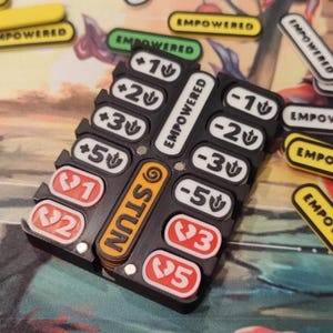 May include: A black plastic board game token organizer holding various rectangular tokens with numerical values and symbols. The text NEW TOKEN EMPOWERED and CHECK THE PERSONALIZATION OPTIONS TO ORDER is visible. Tokens feature plus and minus signs.