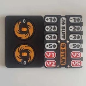 May include: Two black rectangular game tracker boards. The left board features two circular orange dials with a zero indicator. The right board contains white buttons labeled +1, +2, +3, +5, -1, -2, -3, -5, plus red buttons labeled 1, 2, 3, 5, and vertical text reading BUFF and STUN.