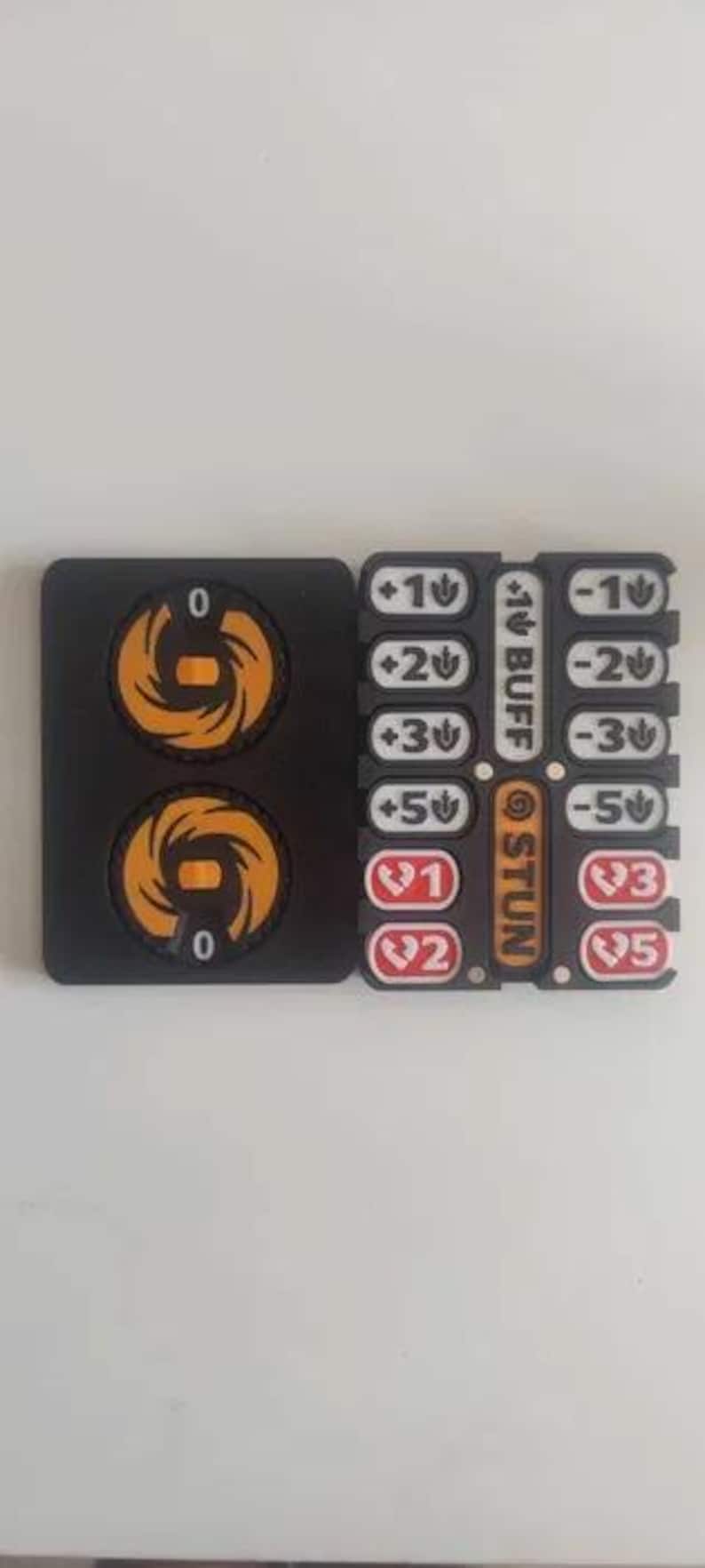 May include: Two black rectangular game tracker boards. The left board features two circular orange dials with a zero indicator. The right board contains white buttons labeled +1, +2, +3, +5, -1, -2, -3, -5, plus red buttons labeled 1, 2, 3, 5, and vertical text reading BUFF and STUN.