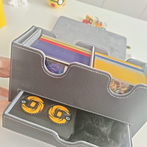 May include: A grey rectangular card storage box with white stitching, featuring two top compartments filled with stacks of colorful trading cards and a bottom pull-out drawer containing two black circular tokens with yellow spiral designs.