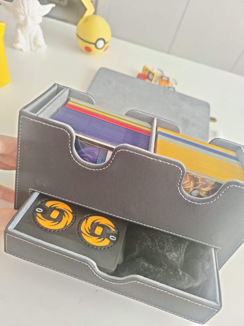 May include: A grey rectangular card storage box with white stitching, featuring two top compartments filled with stacks of colorful trading cards and a bottom pull-out drawer containing two black circular tokens with yellow spiral designs.