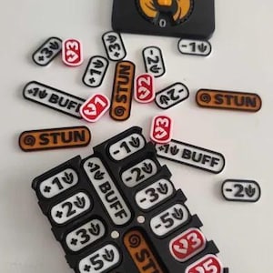 May include: A black plastic game tracker board with two circular orange dials, accompanied by various small rectangular tokens. The tokens feature white, red, and orange backgrounds with printed text including STUN, BUFF, and numerical values.