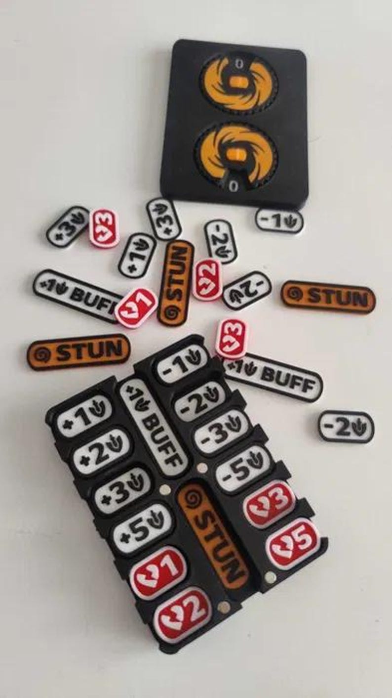 May include: A black plastic game tracker board with two circular orange dials, accompanied by various small rectangular tokens. The tokens feature white, red, and orange backgrounds with printed text including STUN, BUFF, and numerical values.